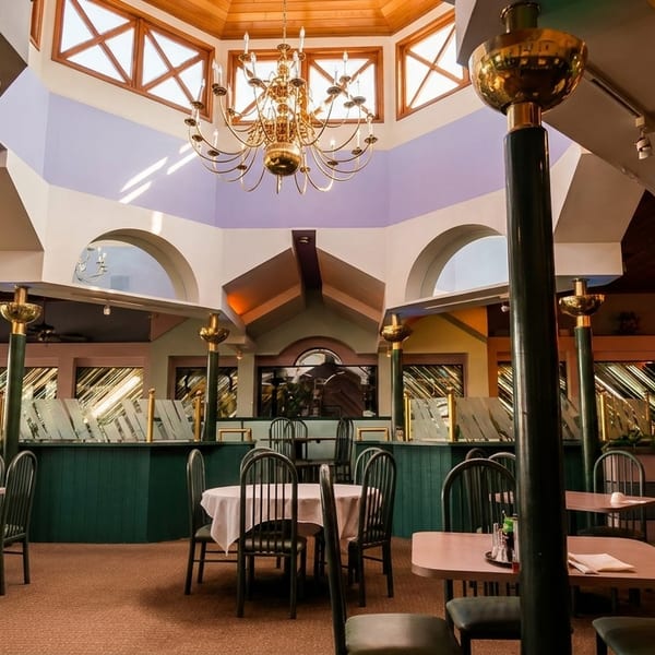 Spacious restaurant dining room with a high ceiling, chandelier, green pillars, and empty tables.